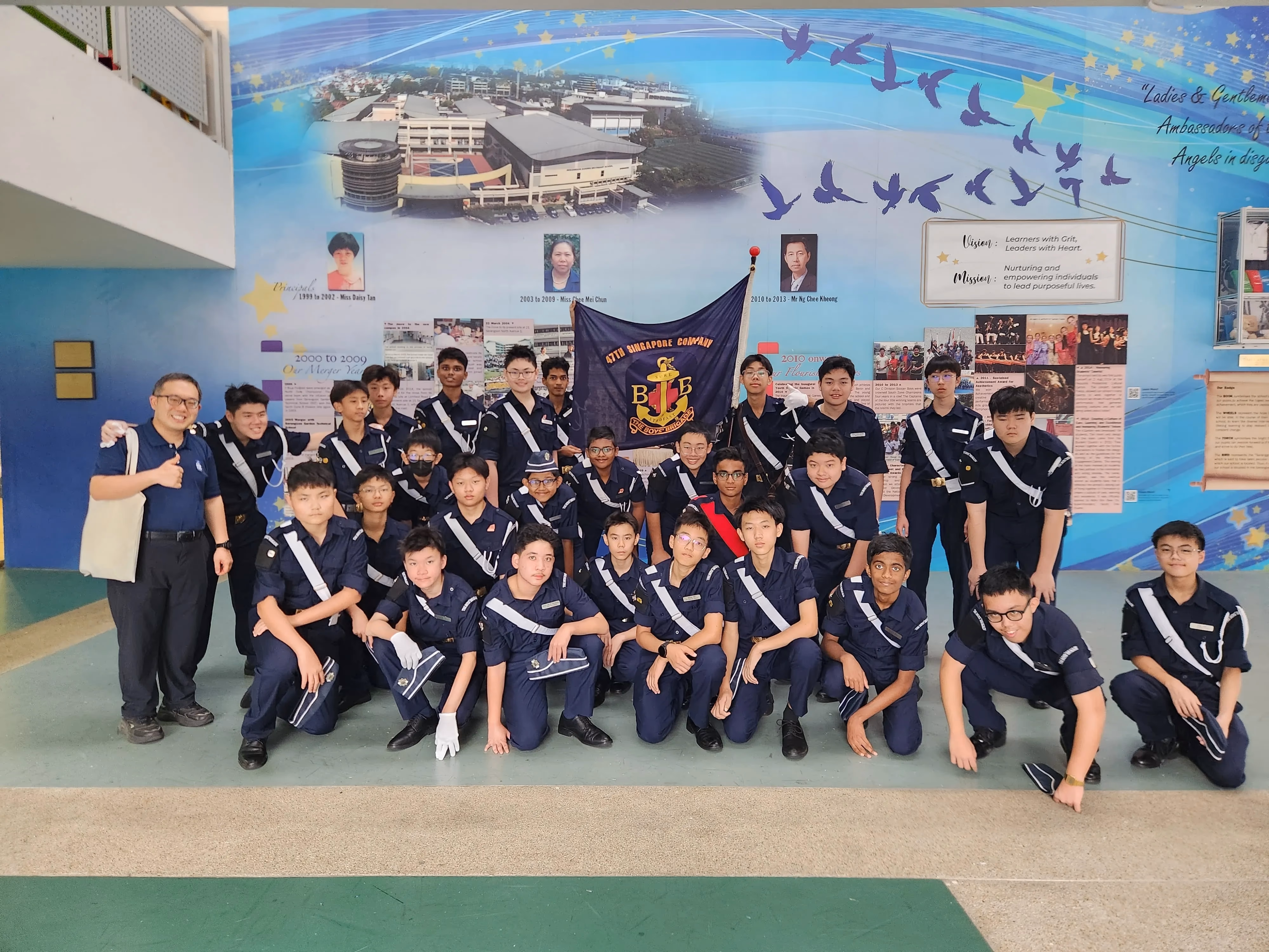 Photograph of a large group of young cadets in navy blue uniforms posing indoors in front of a blue mural featuring birds, a building, and portraits. Group holds a flag with an emblem and text, with a few adults standing on the sides, indicating a formal or commemorative event.